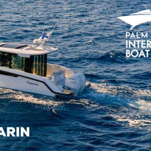 An aerial view of a white Yamarin Aura Cabin motorboat cruising through deep blue ocean water, leaving a white wake behind. The boat features twin outboard engines and a sleek cabin design. The Yamarin logo is in the bottom-left corner, and the Palm Beach International Boat Show logo is in the top-right.