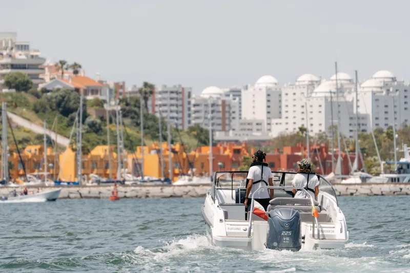 Rear view of a Yamarin 67 DC motorboat cruising away from the camera on a sunny day. Two people are standing in the cockpit, wearing life jackets. The boat is powered by a grey Yamaha V6 outboard motor and is heading toward a coastal city with white and orange buildings in the background.