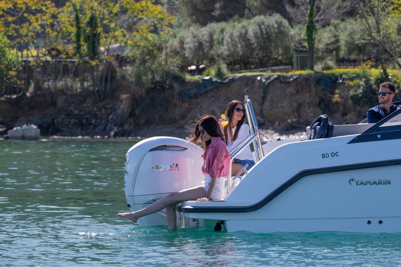 The rear deck and swim platform of a Yamarin 80 DC day cruiser. The boat is equipped with a white Yamaha F350 outboard motor. People are seen enjoying the spacious aft area while the boat is anchored near a sunny coastline.